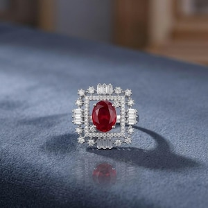 Peut inclure: Une bague argentée ornée d'une grande pierre précieuse rouge foncé de taille ovale au centre. La pierre est entourée d'un cadre rectangulaire incrusté de petites pierres blanches rondes et accentué par quatre pierres blanches taille baguette.
