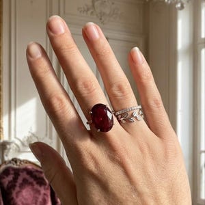 May include: A silver ring featuring a large oval-cut deep red gemstone set in four prongs, accompanied by two stacked silver bands, one with a delicate leaf motif and the other with a row of small clear stones, worn on a finger.