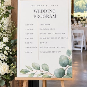 May include: A white wedding program sign on a wooden easel featuring the text OCTOBER 3, 2026, WEDDING PROGRAM, and a schedule of events from 3:00 PM to 9:00 PM. The sign is decorated with a watercolor illustration of green eucalyptus leaves.