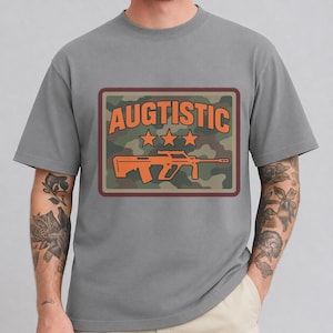 Può includere: Una t-shirt grigia a maniche corte con una grafica rettangolare su sfondo mimetico. La grafica mostra il testo arancione AUGTISTIC sopra tre stelle arancioni e la sagoma arancione di un fucile centrata al centro del rettangolo.