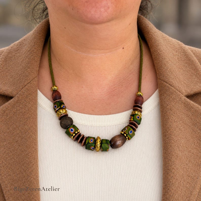 May include: A necklace featuring a green cord strung with a variety of beads, including dark brown wooden beads, textured gold-tone metal spacers, and green cylindrical beads decorated with small blue, yellow, and red circular patterns.