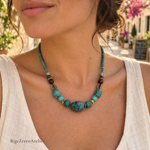 Peut inclure: Un collier de perles avec une grosse pierre centrale de couleur turquoise, entourée de perles turquoise plus petites, de perles sphériques marron foncé et d'entretoises métalliques dorées. Le collier inclut le texte BigeZerenAtelier.