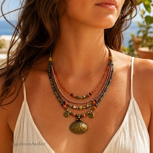 May include: A multi-strand beaded necklace featuring layers of small orange, green, and metallic brown beads. The design includes small circular charms and a large, textured, oval-shaped pendant in a dark bronze tone. The text BigeZerenAtelier is visible.
