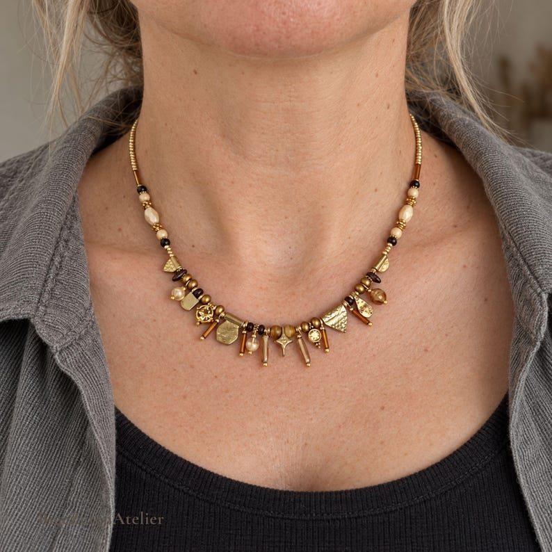 May include: A gold-toned necklace featuring a variety of small charms, including geometric shapes, textured metal pieces, and small beads in shades of cream, brown, and black, arranged along a delicate chain that rests around the neck.