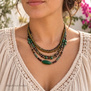 May include: A multi-strand beaded necklace featuring olive green, dark brown, and teal beads. The design includes small round beads, square metallic spacers, and a single elongated oval green bead at the center. The necklace rests against a cream-colored top.