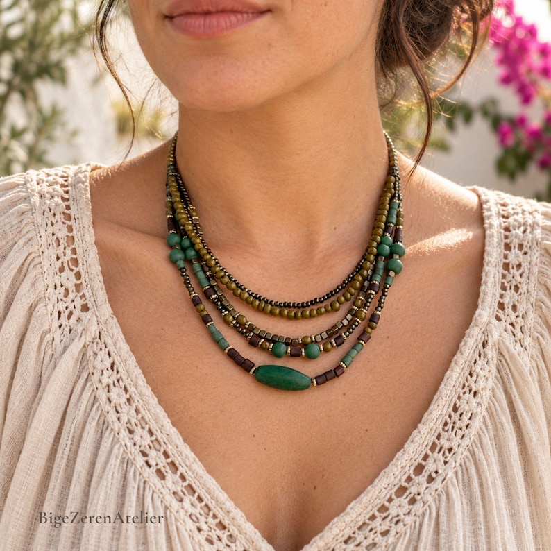 May include: A multi-strand beaded necklace featuring olive green, dark brown, and teal beads. The design includes small round beads, square metallic spacers, and a single elongated oval green bead at the center. The necklace rests against a cream-colored top.