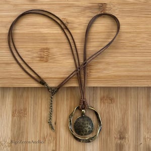 Brown cord pendant necklace laid flat on a wooden background, showing the full necklace, round antique bronze tone pendant, lobster clasp and extension chain.