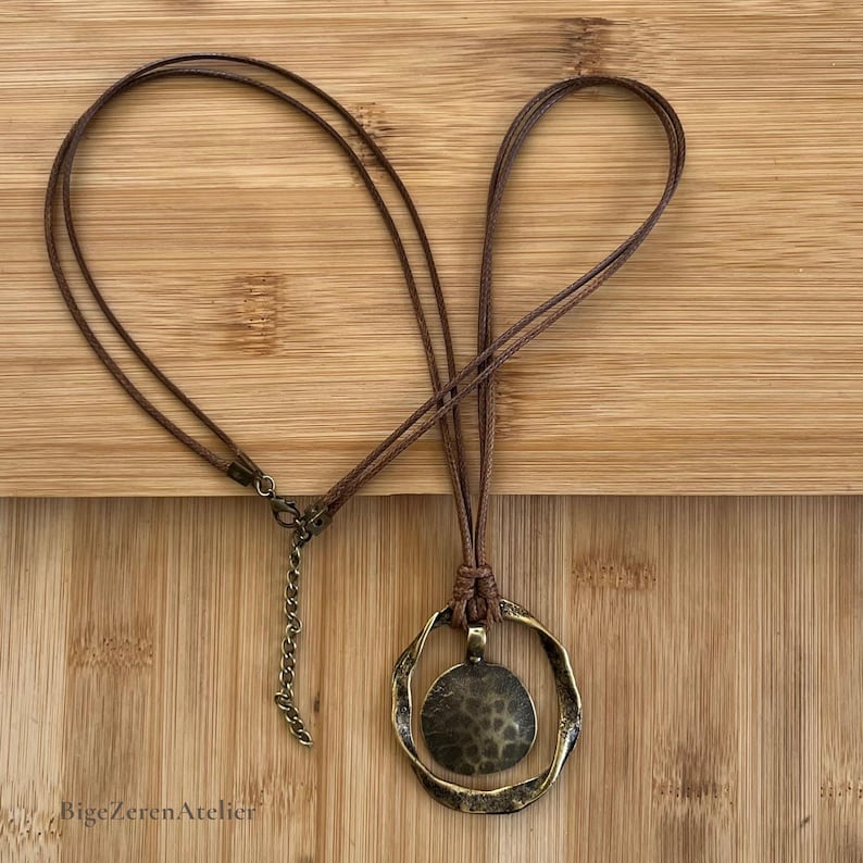 Brown cord pendant necklace laid flat on a wooden background, showing the full necklace, round antique bronze tone pendant, lobster clasp and extension chain.