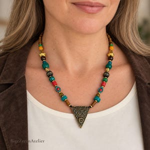 May include: A beaded necklace featuring a central triangular pendant with intricate filigree patterns. The strand consists of multicolored beads in shades of turquoise, red, yellow, brown, and black, accented with metallic gold-toned spacers.
