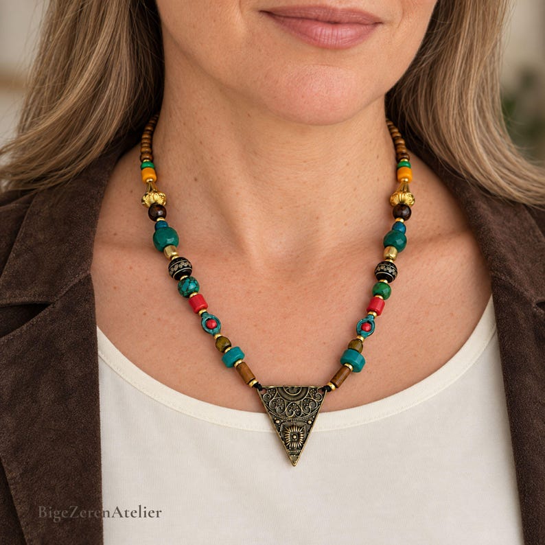 May include: A beaded necklace featuring a central triangular pendant with intricate filigree patterns. The strand consists of multicolored beads in shades of turquoise, red, yellow, brown, and black, accented with metallic gold-toned spacers.