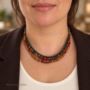May include: A multi-strand beaded necklace featuring layers of dark green, orange, and brown beads. The design includes small metallic spacers, cylindrical wooden beads, and a central, larger, mottled reddish-brown oval bead.