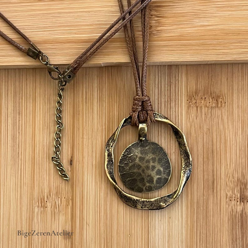 Close-up of the antique bronze tone pendant necklace showing the textured circular center, irregular open frame, brown cord wrapping and adjustable closure.
