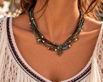 May include: A multi-strand necklace featuring thin dark cords and green threads adorned with small colorful beads in red, yellow, white, and turquoise. A bronze-toned metal maple leaf charm hangs from the center of the layered strands.