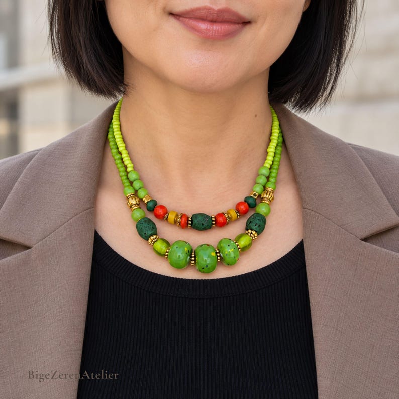 May include: A two-strand necklace featuring lime green seed beads, round orange and yellow beads, and textured dark green beads. The design includes metallic gold-toned spacers between the beads, creating a layered look around the neck.