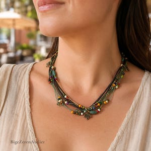 Puede incluir: Un collar de múltiples hilos con cordones finos verdes y marrones adornados con pequeñas cuentas coloridas en tonos rojo, amarillo, azul y blanco. El collar incluye detalles metálicos y un dije central con forma de hoja de arce.