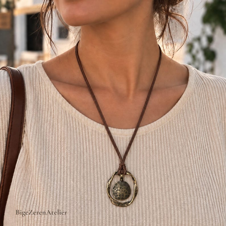 Brown cord pendant necklace worn on a woman, featuring an antique bronze tone irregular round frame and textured circular center pendant.