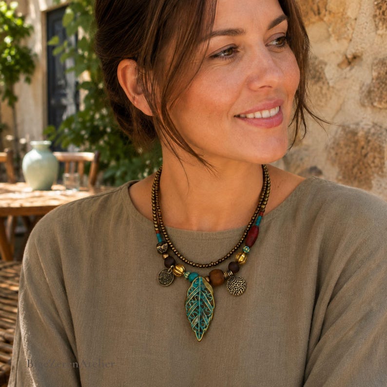 May include: A multi-strand necklace featuring a central leaf-shaped pendant with a teal and bronze finish. The necklace includes small bronze beads, wooden beads, and round metal charms. The design incorporates accents of red and turquoise.