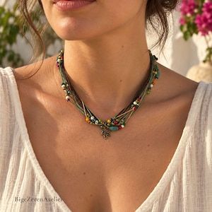 Puede incluir: Un collar de múltiples hilos con cordones finos verdes y marrones adornados con pequeñas cuentas de colores en tonos rojo, azul, amarillo, blanco y verde. Un pequeño dije de metal color bronce con forma de hoja de arce cuelga del centro.
