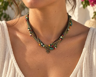 May include: A multi-strand necklace featuring thin green and brown cords adorned with small, colorful beads in shades of red, blue, yellow, white, and green. A small, bronze-toned metal maple leaf charm hangs from the center of the necklace.