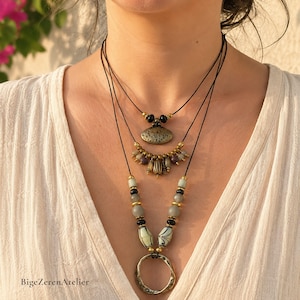 Peut inclure: Un ensemble de colliers superposés composé de trois cordons noirs. Le rang supérieur présente un pendentif ovale en bronze texturé. Le rang intermédiaire comporte de petites perles en bronze et sombres. Le rang inférieur inclut des perles grises, noires et à motifs avec un grand anneau en bronze.