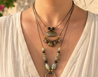 May include: A multi-strand necklace featuring black cords, metallic gold-toned beads, and textured pendants. The design includes a central hammered circular pendant, smaller oval charms, and smooth grey and black beads. The text BigeZerenAtelier is visible.