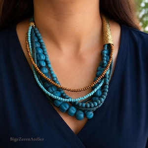 Puede incluir: Un collar de varias hileras con cuentas en tonos azules y turquesas, intercaladas con una hilera de cuentas de color bronce metálico. El collar se sujeta con un cierre cónico de metal dorado texturizado en uno de los lados.