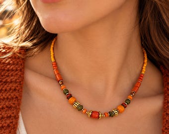 Puede incluir: Un collar de cuentas con una secuencia de cuentas amarillas, naranjas, rojas y verdes intercaladas con pequeños espaciadores de metal dorado y detalles dorados texturizados. El collar incluye el texto BigeZerenAtelier.