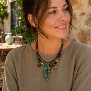 Puede incluir: Un collar de varias hebras con un colgante central en forma de hoja con acabado en bronce y azul verdoso. El diseño incluye pequeñas cuentas de bronce, cuentas de madera y dijes metálicos circulares con patrones texturizados y cuentas turquesas.