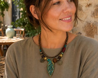 May include: A multi-strand necklace featuring a central teal and bronze-toned leaf pendant, accented with small bronze beads, wooden beads, and circular metal charms. The design incorporates turquoise and red bead accents along the layered chains.