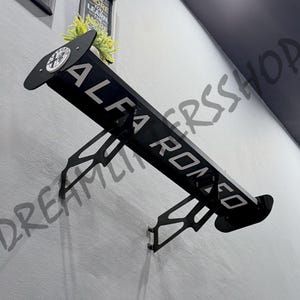 May include: A black automotive rear spoiler mounted to a wall, featuring the white text ALFA ROMEO across the blade and a circular Alfa Romeo logo on the side end plate. The spoiler is supported by two black metal brackets with cut-out designs.