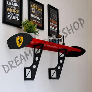 May include: A red wall-mounted shelf designed to resemble a race car spoiler, featuring two black support brackets with circular cutouts and black oval end plates. The left end plate displays a yellow Ferrari logo with a black prancing horse.