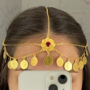May include: A gold-toned headpiece featuring a central floral-shaped ornament with a red stone, connected to a delicate chain that runs along the forehead. The piece is adorned with multiple small, circular gold-toned coin charms hanging down.
