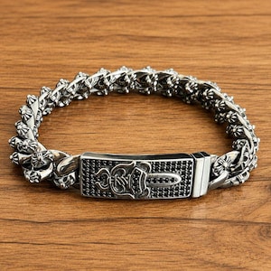 May include: A silver-toned chain link bracelet featuring a rectangular clasp adorned with black stones and a central raised dagger emblem. The bracelet links are detailed with small cross-shaped motifs throughout the entire length of the piece.