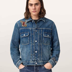 May include: A medium-wash blue denim jacket featuring a pointed collar, metal button closures, and two chest pockets. An embroidered eagle patch is on the right chest, and the letters AS are embroidered in black on the left sleeve cuff.