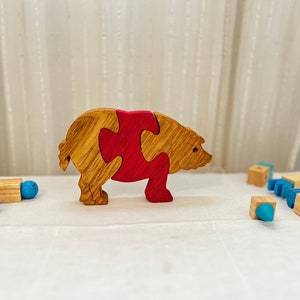 May include: A wooden puzzle toy shaped like a pig. The pig is made of two pieces, one brown and one red, that fit together. The pig is standing on a white surface.