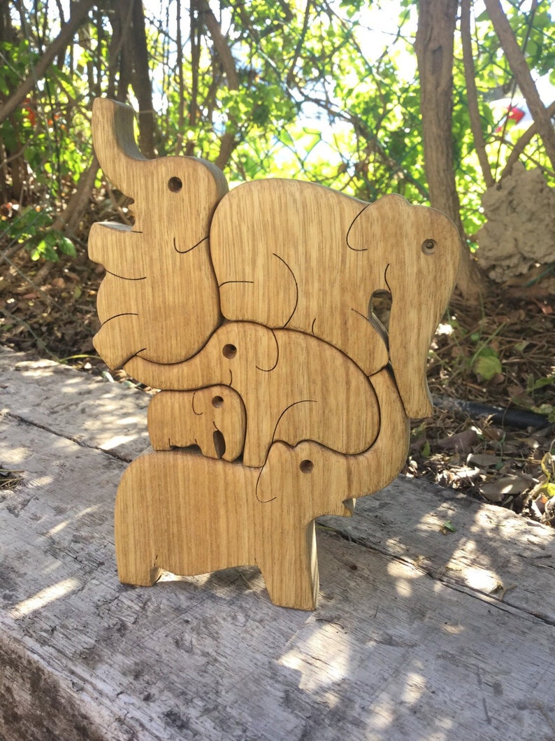 Handcrafted wood elephant puzzle displayed in its original stacked design, with five interlocking elephant shapes of varying sizes made from natural African Walnut, set against a rustic outdoor backdrop with greenery and sunlight through the leaves.