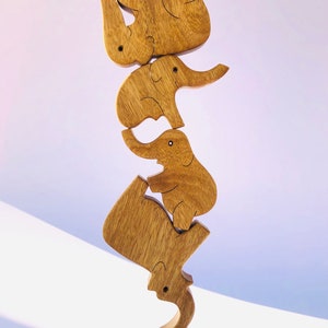 The wooden elephant family puzzle displayed in a vertical, gravity-defying balance. Each elephant piece, from the largest to the smallest, is stacked in a playful, ascending tower on a soft pink backdrop.