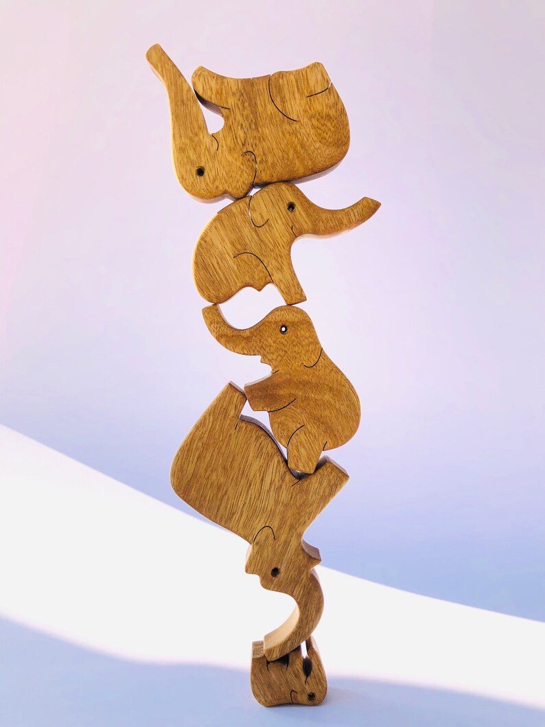 The wooden elephant family puzzle displayed in a vertical, gravity-defying balance. Each elephant piece, from the largest to the smallest, is stacked in a playful, ascending tower on a soft pink backdrop.