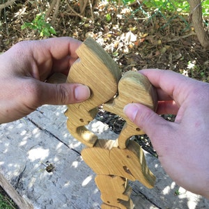 A pair of hands carefully balancing the wooden elephant puzzle pieces on a log outdoors, demonstrating the game aspect of the set with green foliage in the background.