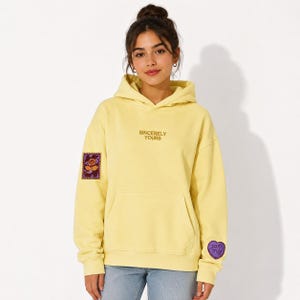 Puede incluir: Una sudadera con capucha de color amarillo pálido con el texto SINCERELY YOURS bordado en marrón en el pecho. En la manga derecha hay un parche rectangular con un gráfico de rosa y en la izquierda un parche de corazón morado con el texto yours truly.