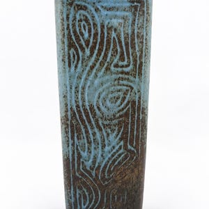 May include: A tall, rectangular ceramic vase featuring a dark brown base glaze with a textured, light blue overlay. The surface is decorated with carved, swirling, and linear patterns that reveal the dark brown clay beneath the blue glaze.