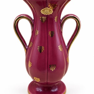 May include: A magenta porcelain vase with a scalloped rim, two curved handles, and a gold-painted shell pattern. The base and rim are trimmed in gold. A gold sticker near the top reads Gefle Porslin with a logo. The body has vertical fluting.