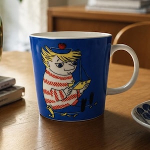 Peut inclure: Un mug en céramique bleu orné d'une illustration graphique représentant un personnage blond vêtu d'un haut à rayures rouges et blanches et d'un chapeau bleu, tenant un petit bateau jaune. Le mug possède un intérieur et une anse blancs.