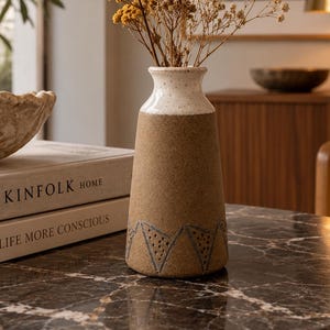 Könnte beinhalten: Eine Keramikvase mit gesprenkeltem weißem Hals und einem strukturierten beigen Körper, der an der Basis ein sich wiederholendes Dreiecksmuster aufweist. Die Vase enthält einen Strauß aus getrockneten gelben Schafgarben und weißen Schleierkrautblüten.