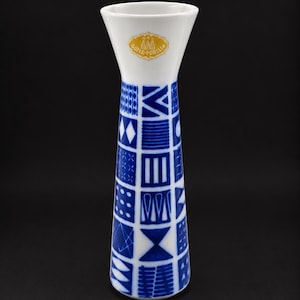 Pode incluir: Um vaso de porcelana branca com borda alargada e corpo cônico, decorado com uma grade de padrões geométricos azuis. Um adesivo dourado perto do topo traz a inscrição GÜTEE-PORSLIN com um pequeno ícone de edifício acima do texto.