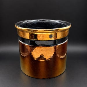 May include: A cylindrical ceramic planter featuring a reflective metallic copper finish on the lower body, a wide horizontal band of glossy black glaze in the centre, and a polished gold-toned rim at the top with a dark interior finish.