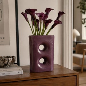 Può includere: Un vaso rettangolare in ceramica viola con due aperture circolari al centro, contenente un bouquet di calle viola scuro. Il vaso presenta una finitura lucida e una forma organica leggermente irregolare posizionato su una superficie in legno.