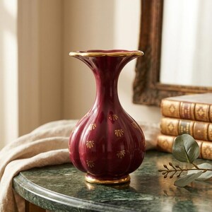 May include: A deep burgundy ceramic vase with a fluted, bulbous body and a flared rim. The vase features a glossy finish, gold-painted shell-like motifs scattered across the surface, and gold trim along the top edge and the circular base.