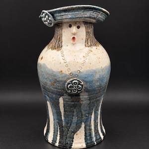 Op de afbeelding: Een keramische kan in de vorm van een persoon met een hoed met brede rand. Het figuur heeft een crèmekleurig gezicht met bruin haar, een kleine open mond en een blauw-wit gespikkeld lichaam met een ronde hanger en verticale blauwe strepen.