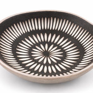 May include: A shallow, round ceramic bowl with a matte black interior featuring a white radial pattern of elongated, petal-like shapes. The exterior rim and base of the bowl are finished in a smooth, light beige, contrasting with the dark interior.